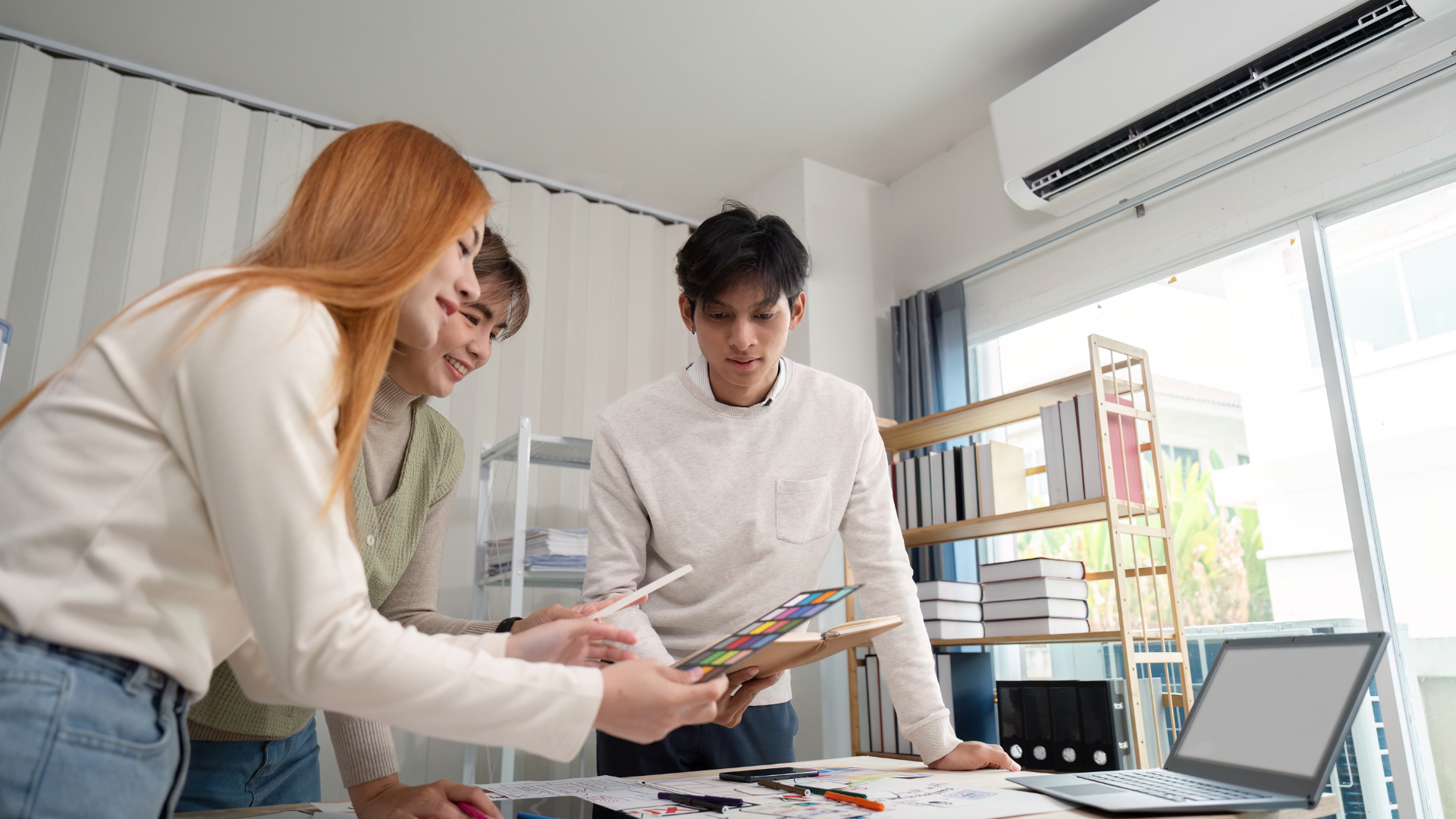 Three young professionals collaborating in a modern office, reviewing color samples and design sketches on a desk.