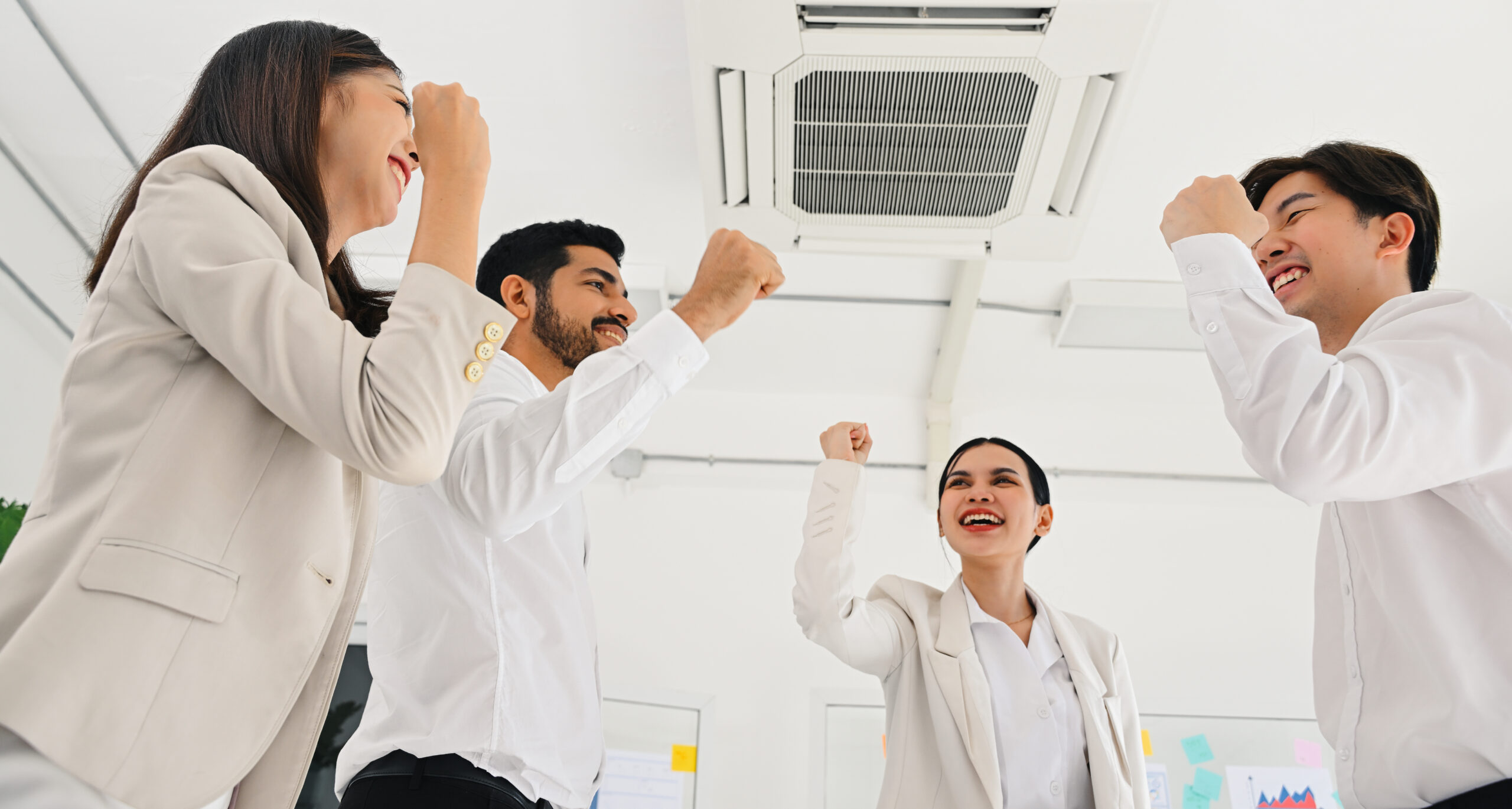 Group of smiling business professionals raising their fists in celebration under a ceiling-mounted air conditioner in a bright office.