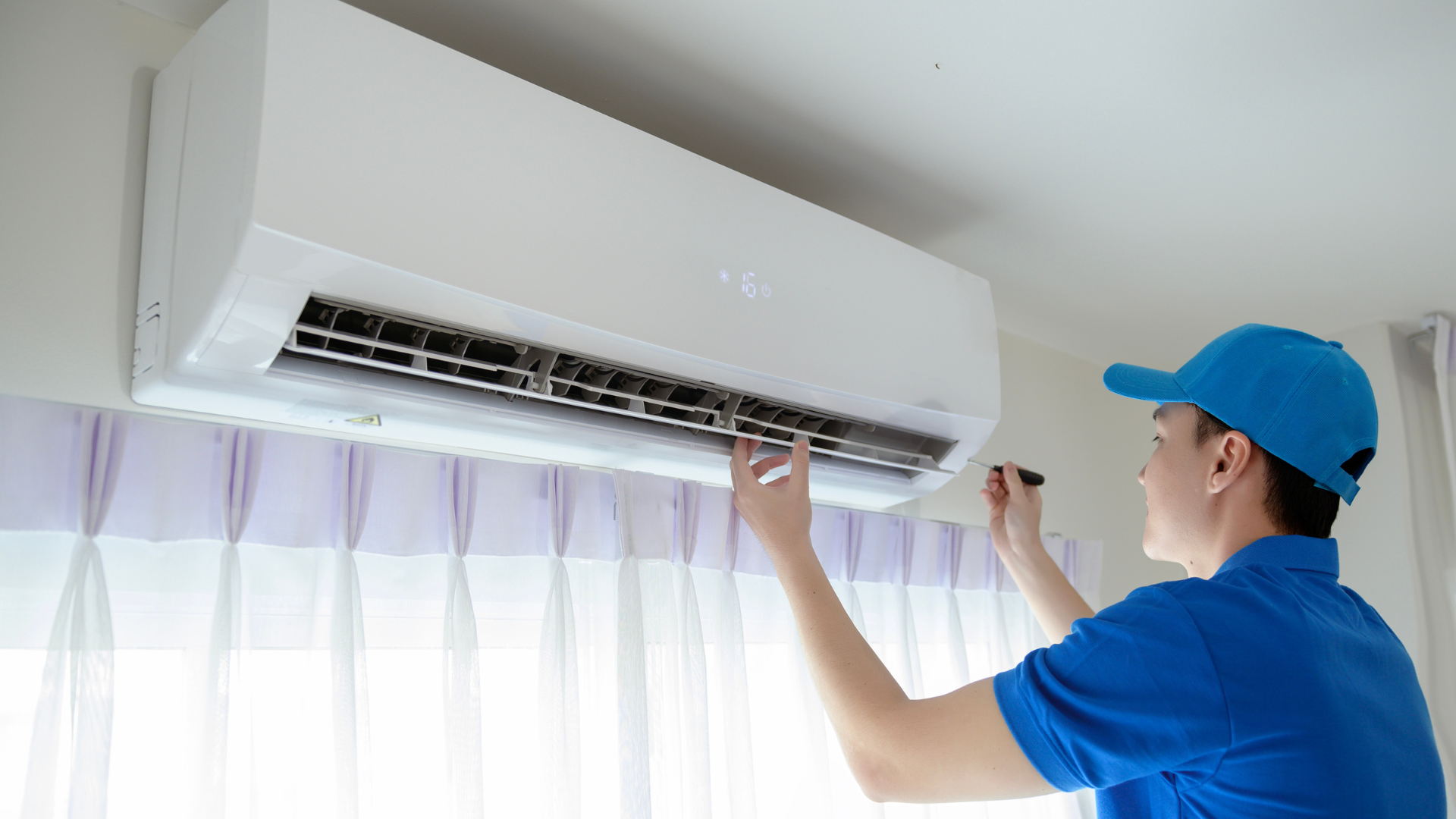 A technician in a blue uniform and cap performing maintenance on a white wall-mounted air conditioning unit inside a home.