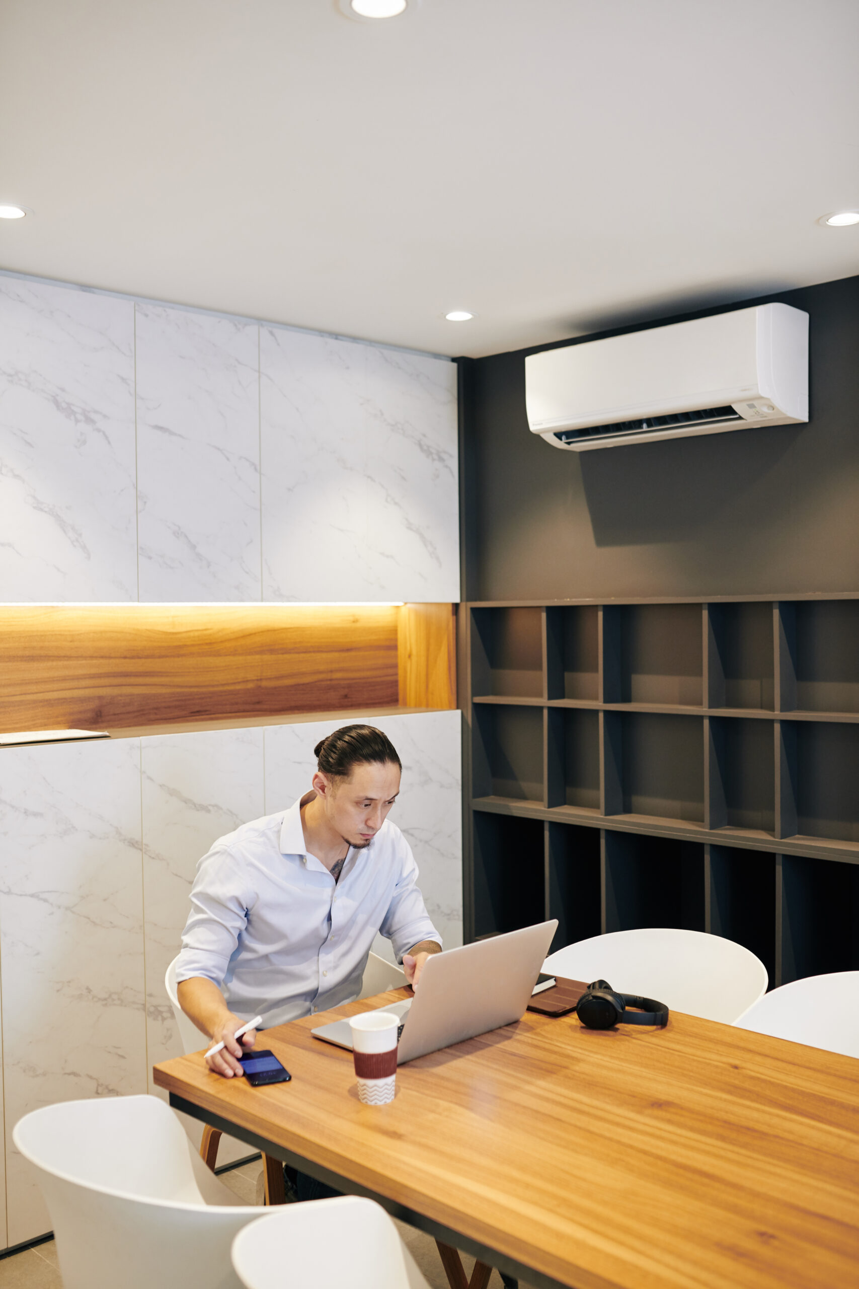 Man working on a laptop at a wooden desk in a modern office with wall-mounted air conditioning unit.