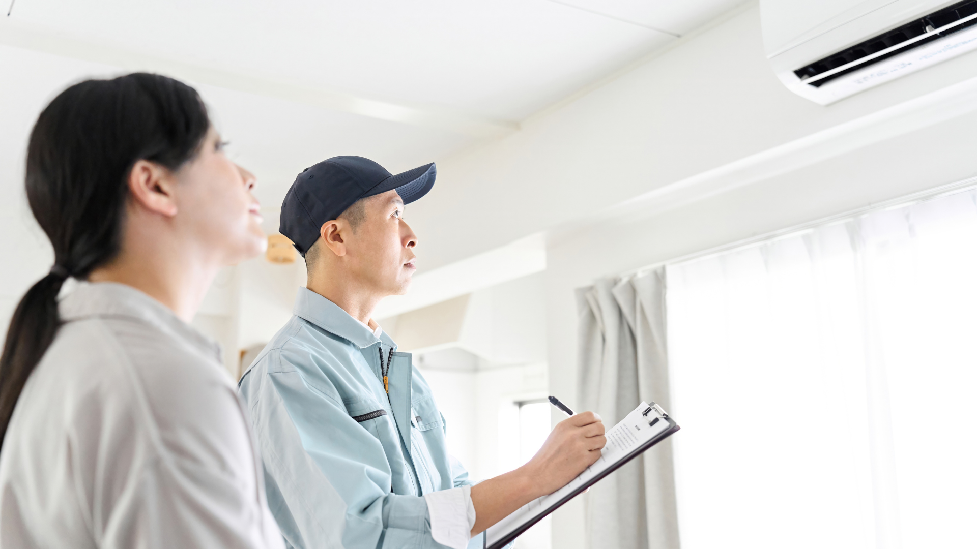 A technician in a light blue uniform and cap writes on a clipboard while inspecting an air conditioning unit on the ceiling, with a homeowner observing nearby.