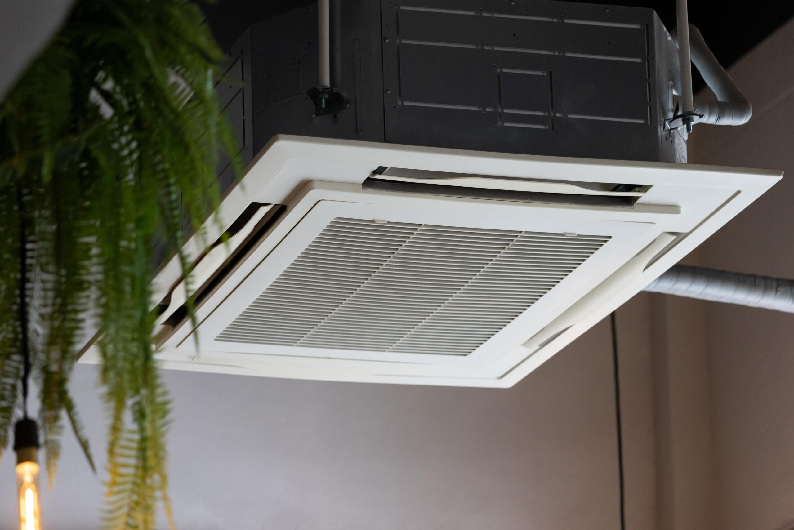 White ceiling-mounted cassette air conditioner with central grille and square panel design installed in commercial interior with exposed ductwork and hanging plants
