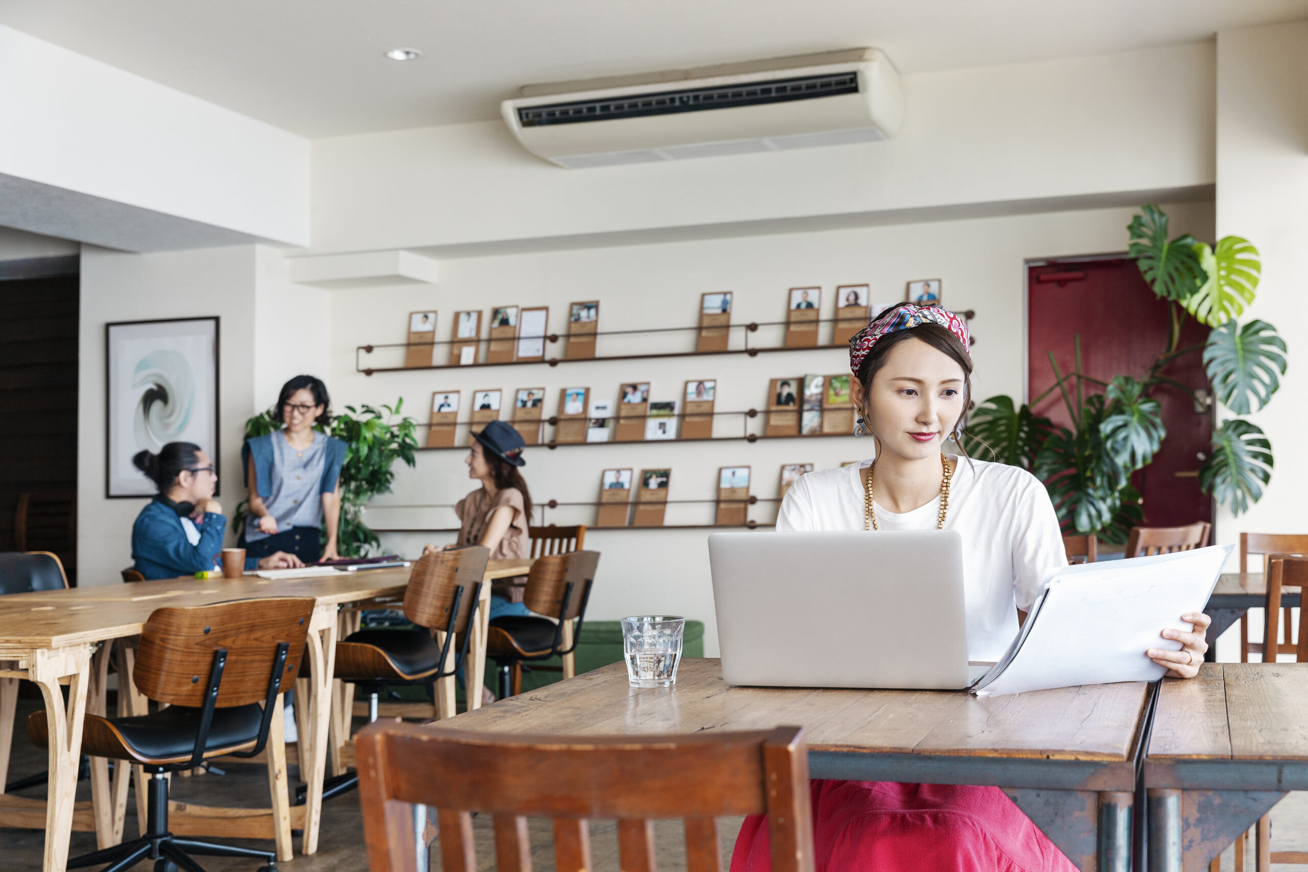 Modern cafe interior with ceiling-mounted air conditioning unit, woman working on laptop at wooden table, customers in background, wall-mounted display shelves, and tropical plants creating comfortable workspace atmosphere